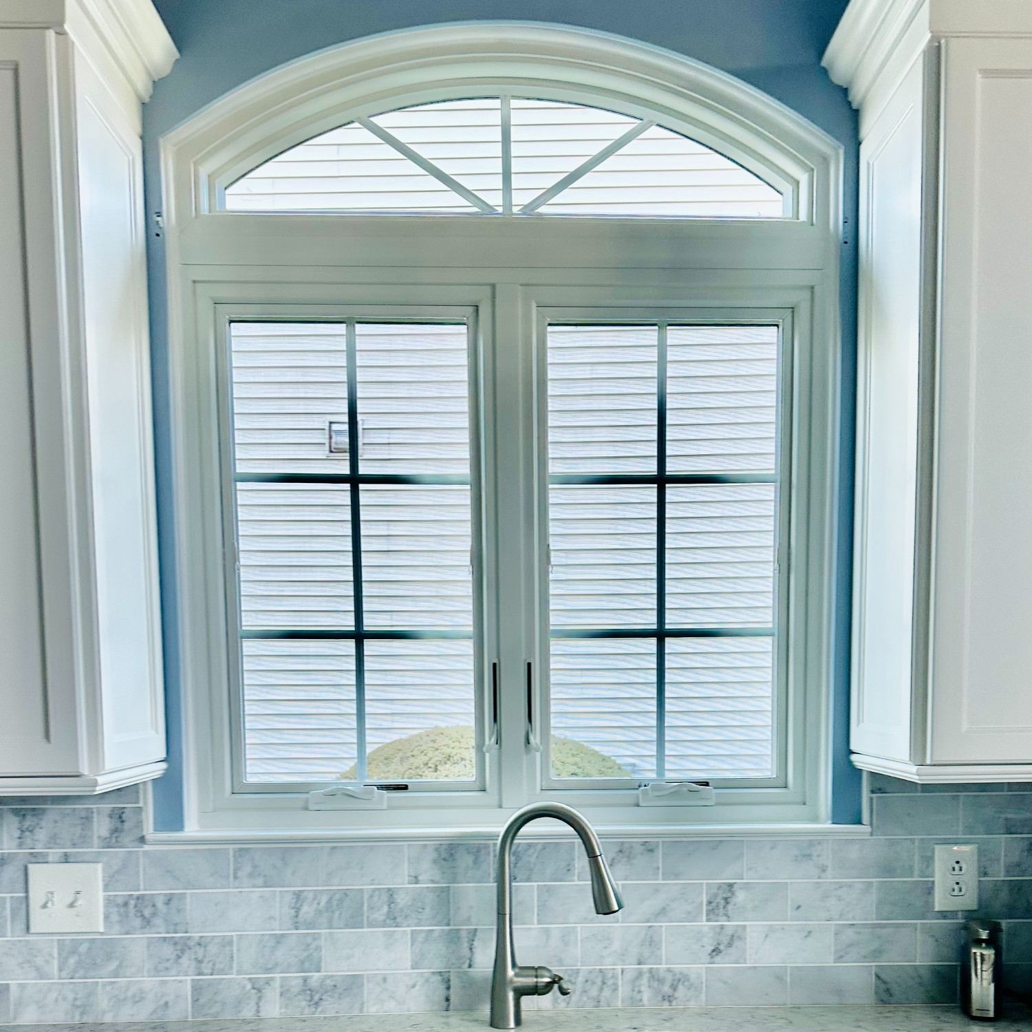 An equal leg arch with sunburst grilles above two casements. The window is white and in a kitchen above sink and between white cabinets.