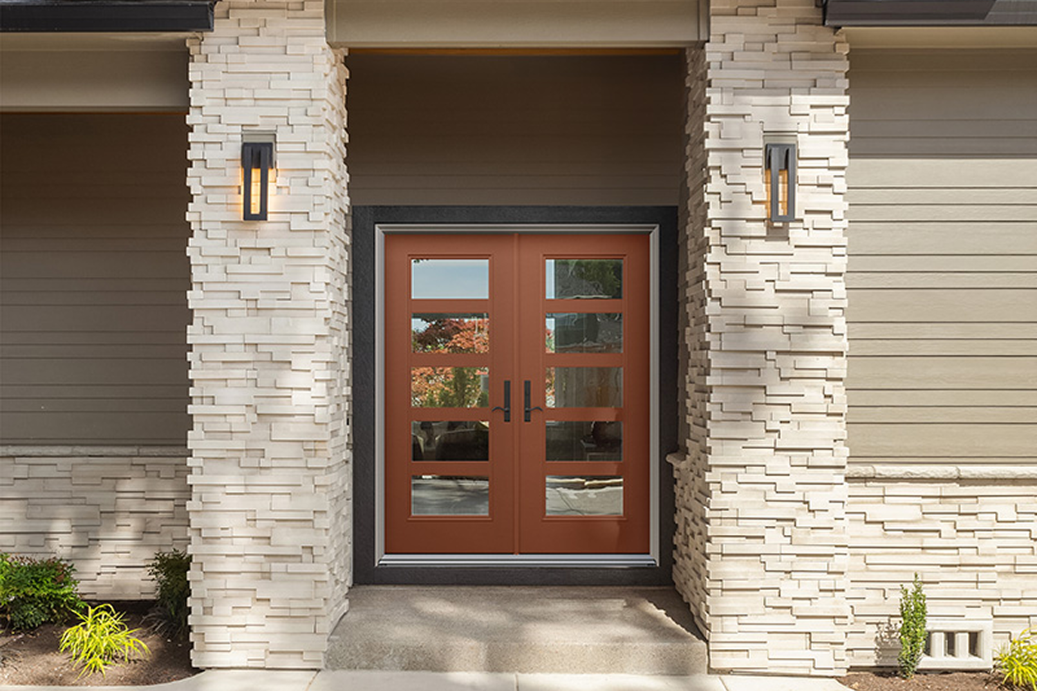 Double entry door made of composite wood with 4 horizontal glass sections in each door. Two tan stone columns sit further in front of the door and frame the entrance to the home.